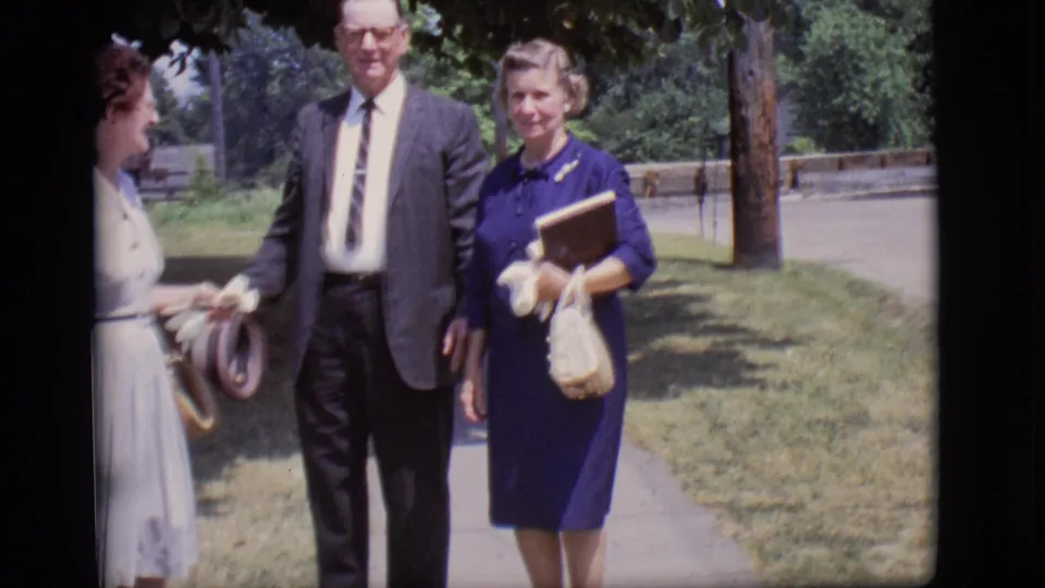 In 1966 Des Moines home-movie footage, a suited man walks with two women, one in a light dress and one in a blue dress carrying a book and purse.
