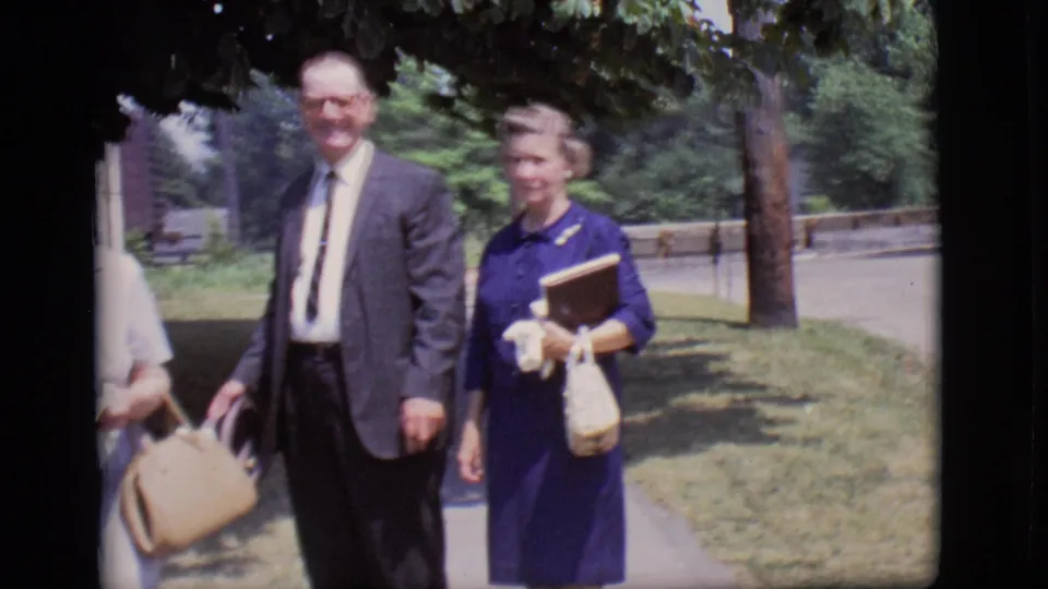 A home-movie frame from 1966 Des Moines shows the adults walking past the camera in formal clothing, with a sidewalk and trees behind them.