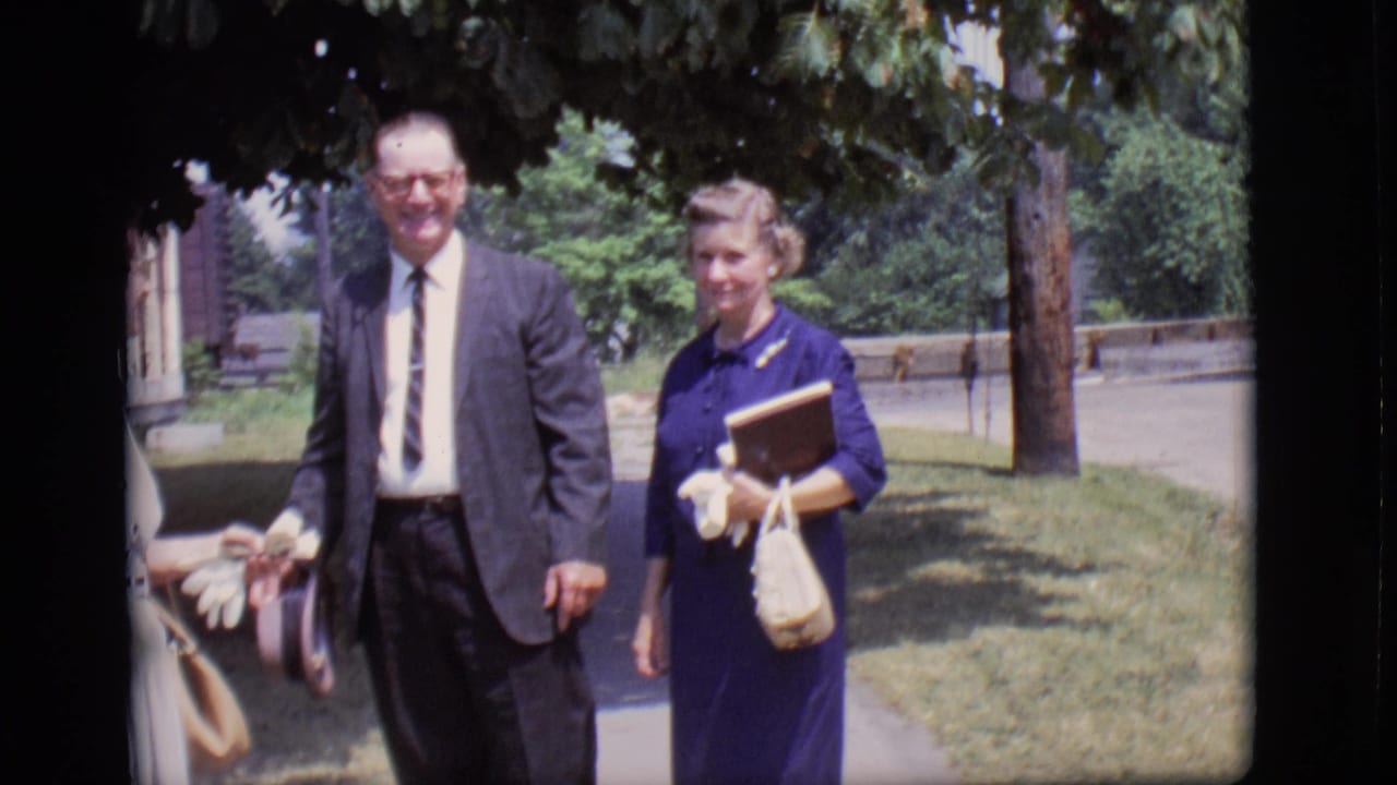 Older man in a suit walks beside a woman in a blue dress on a tree-lined sidewalk.