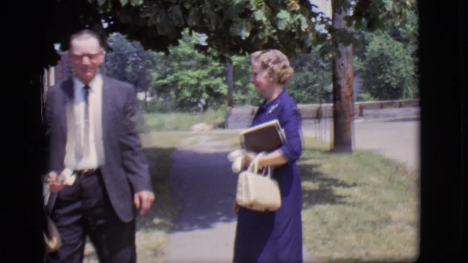 A suited older man moves past the camera as a woman in a blue dress holds a book and purse near the church sidewalk.