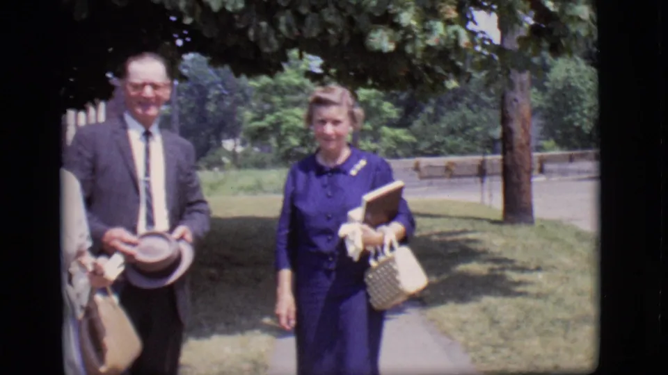 An older man with a hat and a woman in a blue dress walk toward the camera on a sunny Des Moines sidewalk in 1966.