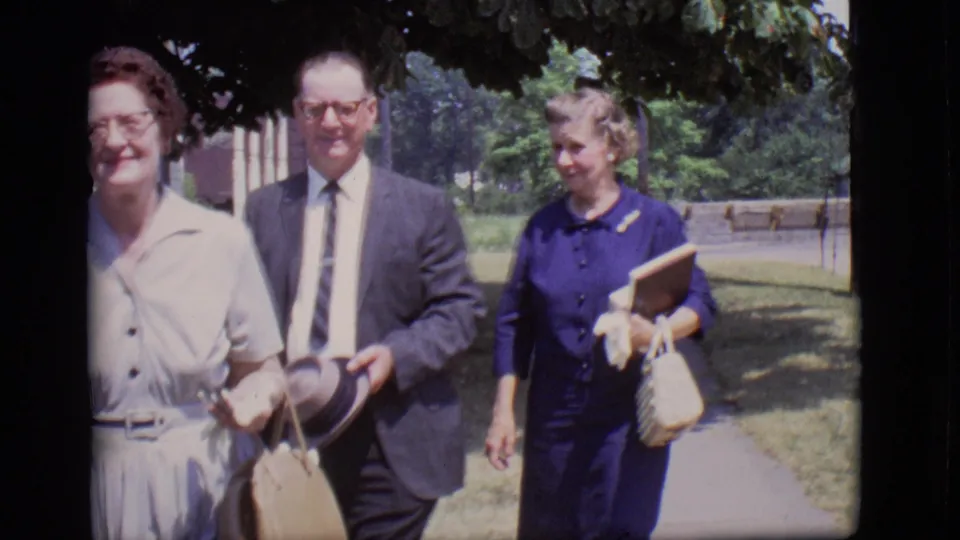 Three older adults, including a woman in a light dress, a suited man, and a woman in blue, walk along the shaded sidewalk.