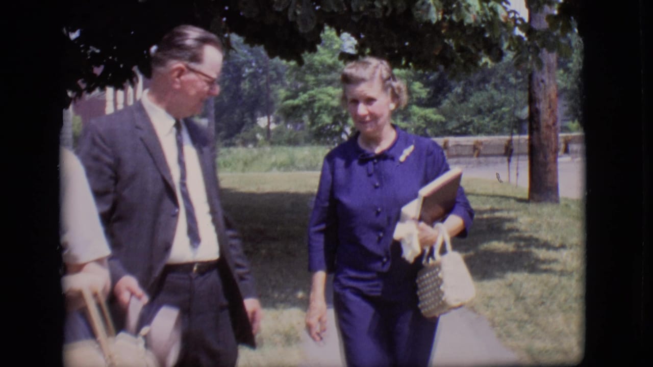Man in a dark suit walks beside a woman in a purple dress on a tree-lined path.