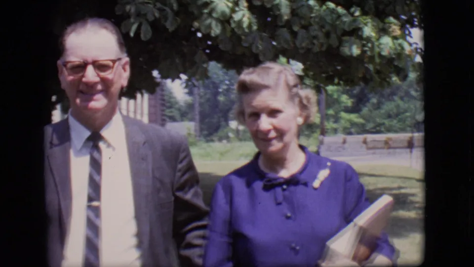 A closer 1966 Des Moines frame shows the man in glasses and tie beside the woman in a purple dress holding a book or folder.