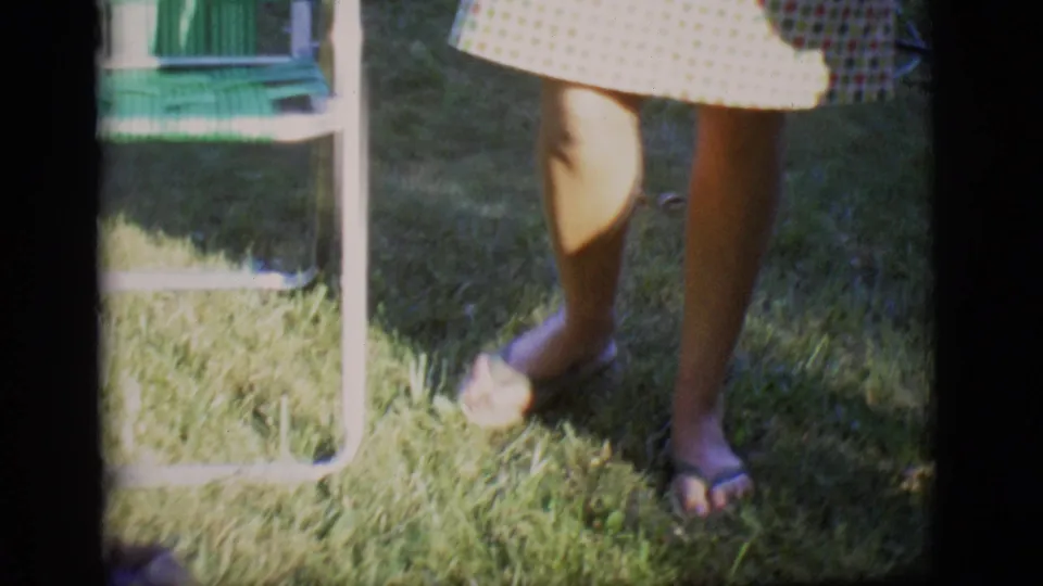 A 1966 backyard frame centers on the adult's legs, checkered dress hem, flip-flops, grass, and the nearby woven lawn chair.