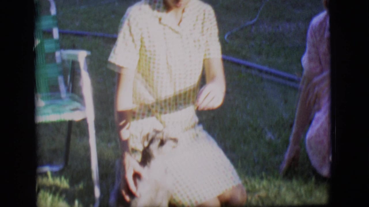 A woman in a yellow checkered dress interacts with a small dog on a sunlit lawn in the Des Moines home-movie clip.