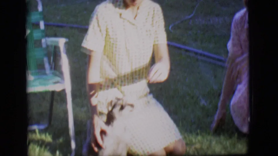 A woman in a yellow checkered dress interacts with a small dog on a sunlit lawn in the Des Moines home-movie clip.