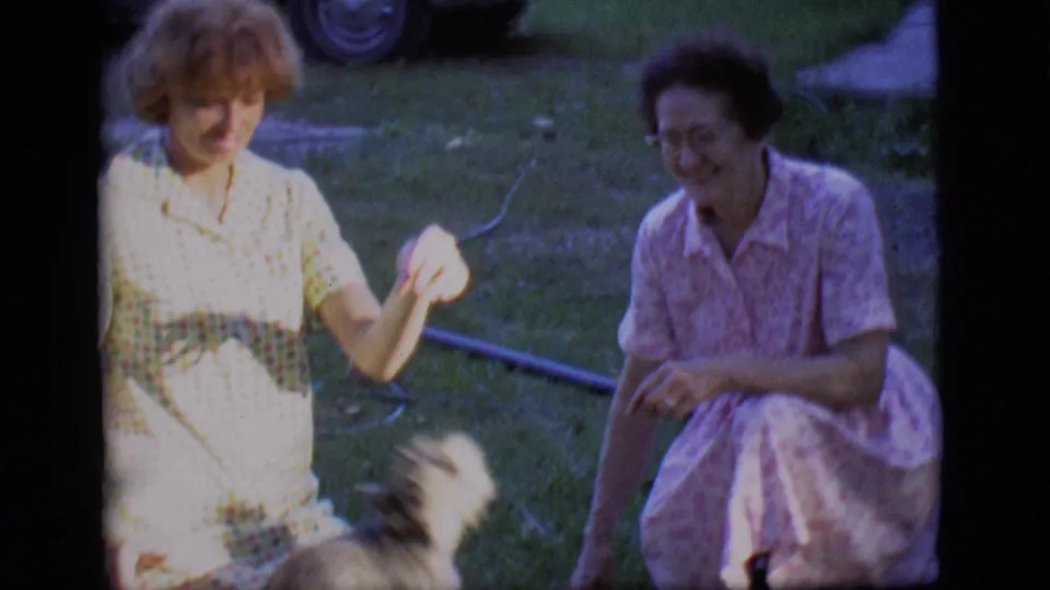 A woman gestures toward the small dog at the edge of frame while a second woman crouches smiling on the backyard lawn.
