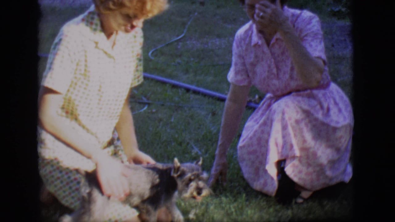 In 1966 Des Moines home-movie footage, two women in patterned dresses kneel and sit on grass while petting a small dog.