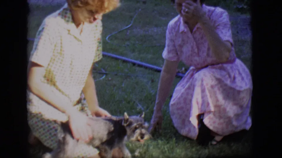 In 1966 Des Moines home-movie footage, two women in patterned dresses kneel and sit on grass while petting a small dog.