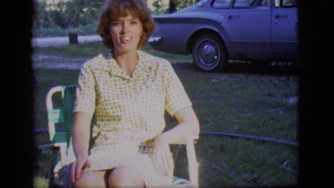 Woman seated in a green folding chair with a blue sedan behind her.