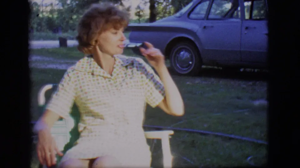 A woman seated on a lawn chair turns in profile during a 1966 outdoor home-movie scene, with a blue sedan in the background.