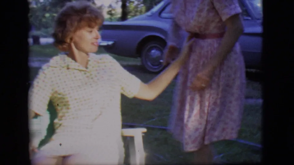 On a grassy lawn in Des Moines, one woman sits in a chair and reaches toward another woman standing nearby, with a blue sedan behind them.