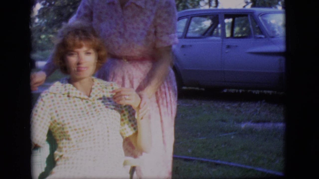 Younger woman seated on a lawn while an older woman stands behind her near a parked sedan.