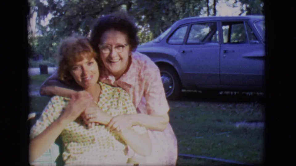 In this 1966 Des Moines frame, two adult women pose closely on a lawn as the older woman embraces the seated younger woman from behind.