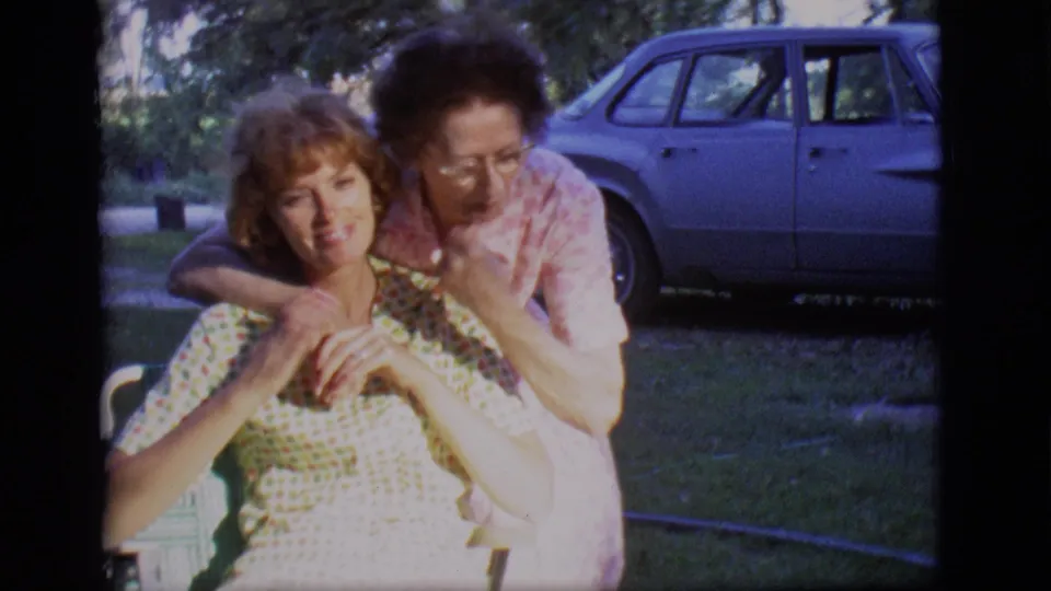 A 1966 Des Moines home-movie frame shows an older woman leaning over a seated younger woman, their hands together, with a sedan behind them.