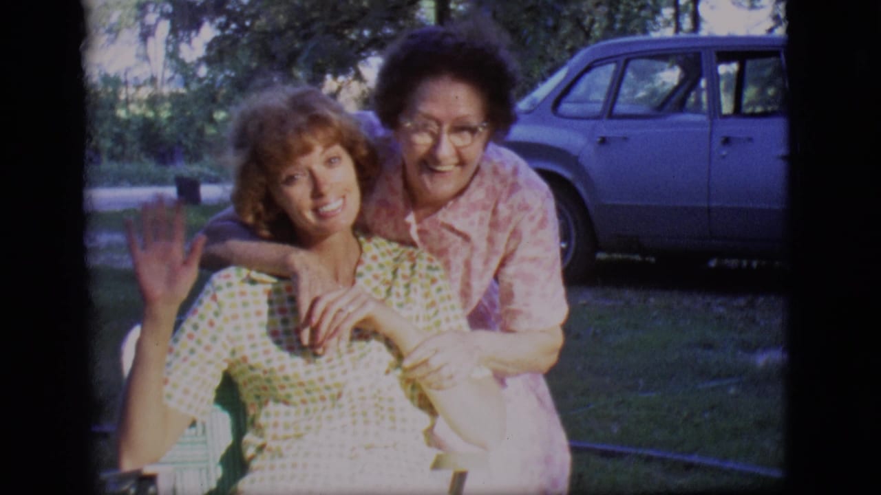 Two women smiling in a backyard, with one seated and one leaning over her shoulder.