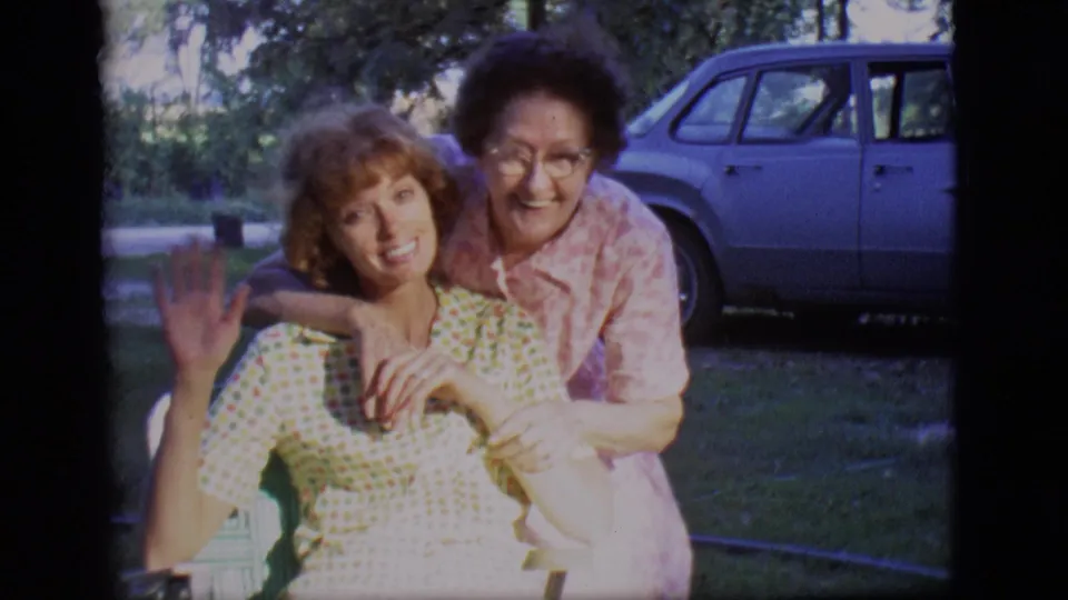 In 1966 Des Moines home-movie footage, two women smile in a grassy backyard as one waves beside a parked sedan.