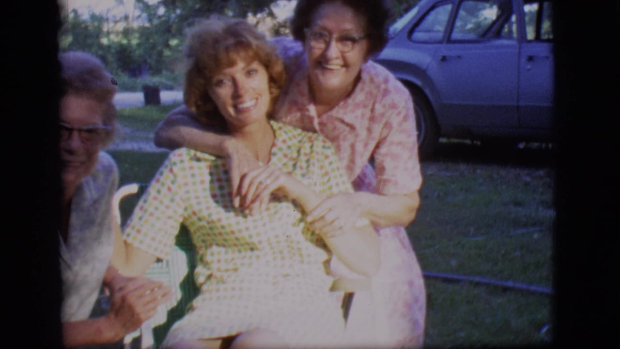 Three women gather around a chair in a backyard, smiling toward the camera.