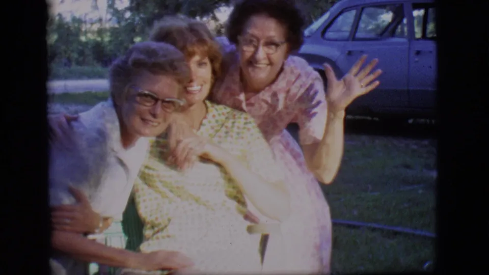 Three women in 1966 Des Moines footage lean together, wave, and smile in a bright backyard scene.