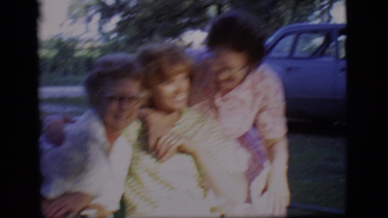 Three women sit close together in a front yard, with a blue car behind them.