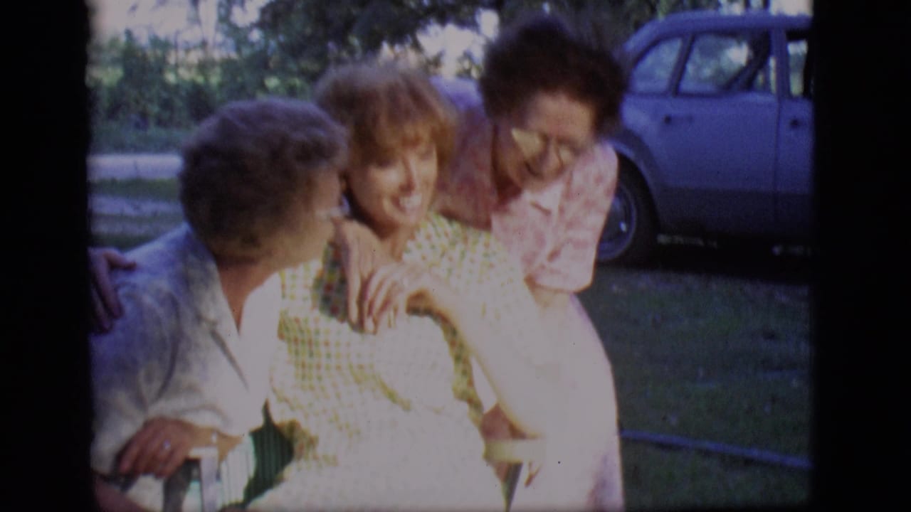 Two women lean toward a seated younger woman in a lawn chair outdoors.