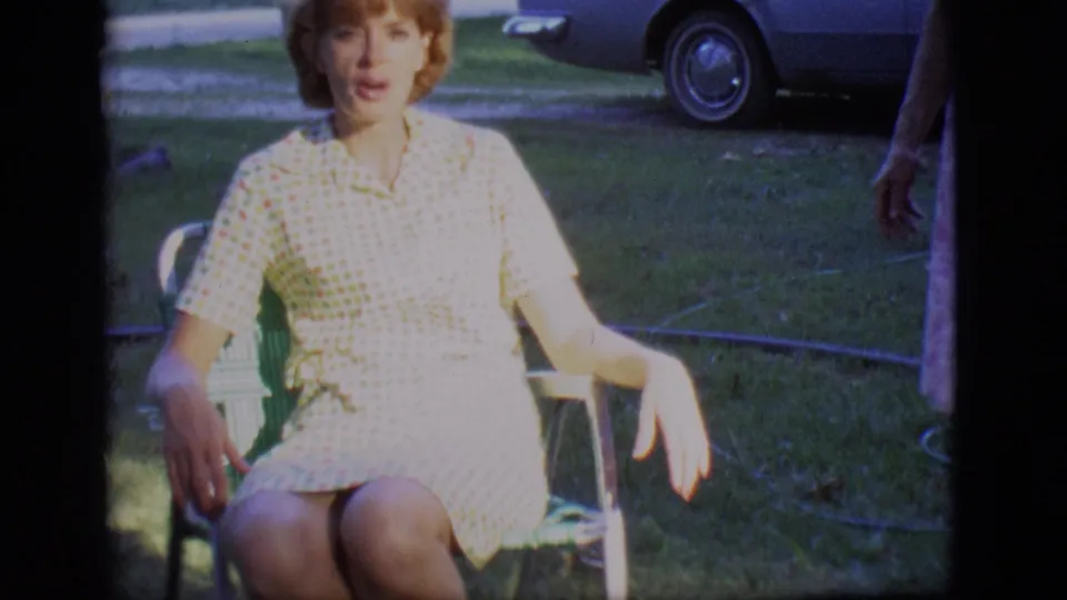 A younger woman in a checkered dress sits alone in a lawn chair in the Des Moines yard, with grass and a parked car behind her.