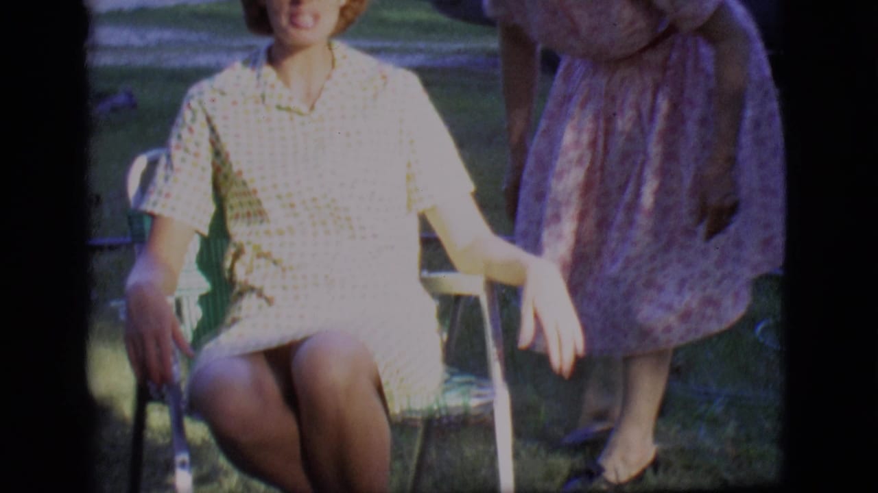 Seated woman in a checkered dress rests her arms on a lawn chair beside a standing woman.