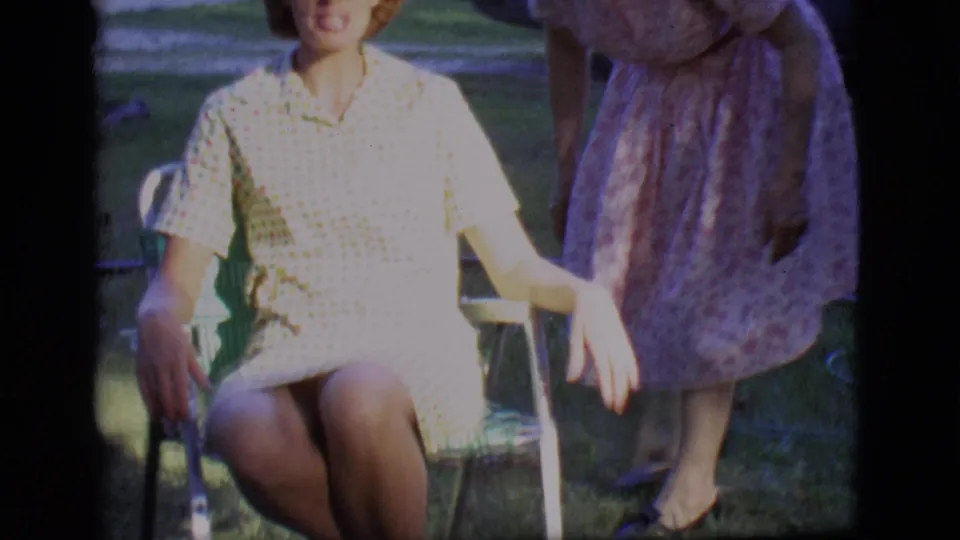 A 1966 backyard frame shows the seated woman in a pale checkered dress with a companion standing close by on the grass.