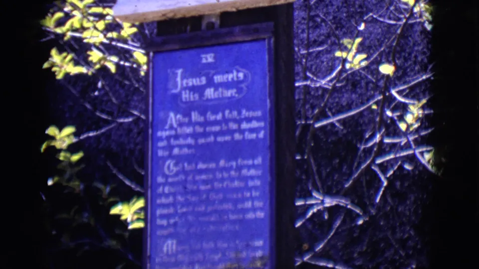 1965 San Luis, Colorado frame showing a blue Stations of the Cross plaque against stone, partly framed by branches and bright leaves.