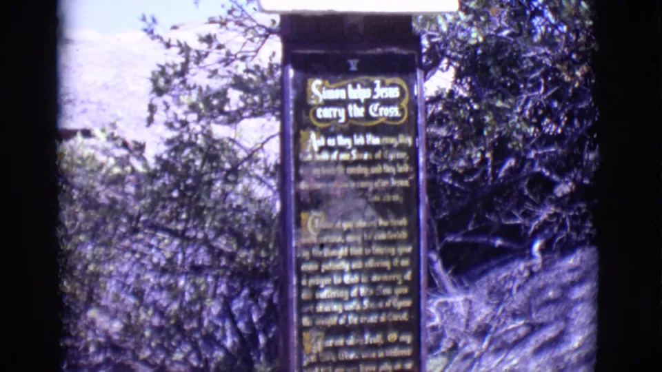 1965 San Luis, Colorado frame with a dark station sign titled Simon helps Jesus carry the Cross, partly framed by brush and trees.
