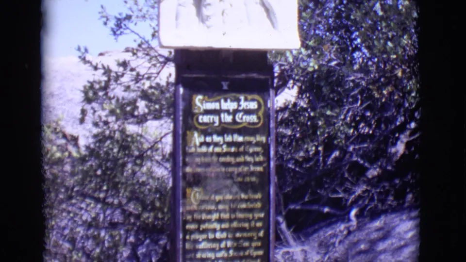 1965 San Luis, Colorado 8mm frame showing the Simon helps Jesus carry the Cross sign beneath a pale relief panel near trees.