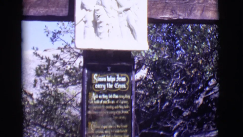 1965 San Luis, Colorado frame of the Simon helps Jesus carry the Cross station with a carved relief above and dense foliage behind.