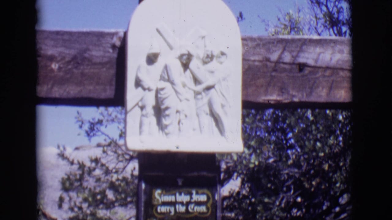 White relief sculpture above a plaque on a wooden cross outdoors.