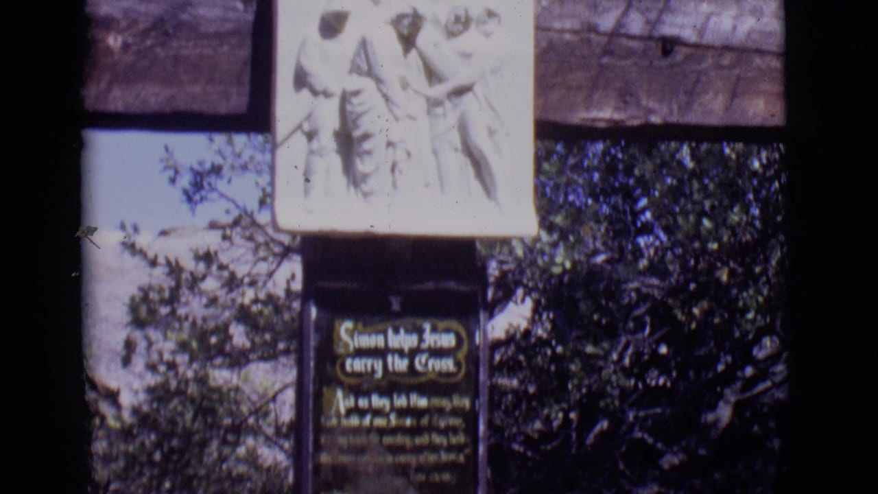 San Luis, Colorado home-movie frame of a Christian station with relief figures, a titled plaque, wooden supports, and surrounding trees.