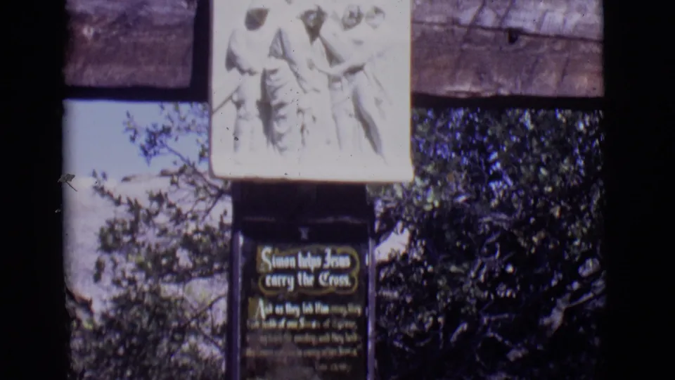 San Luis, Colorado home-movie frame of a Christian station with relief figures, a titled plaque, wooden supports, and surrounding trees.