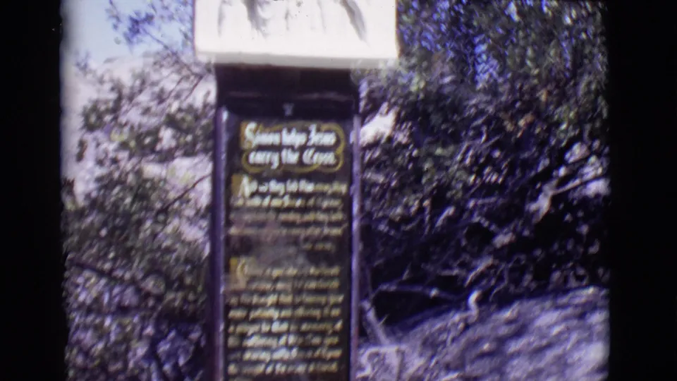8mm frame from San Luis, Colorado showing the lower plaque and relief panel of the Simon and Jesus cross-carrying station amid trees.