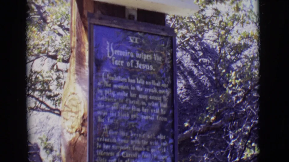 Close 1965 8mm view of a blue Christian station plaque referencing Jesus, with a carved wooden support and leafy trees behind it.