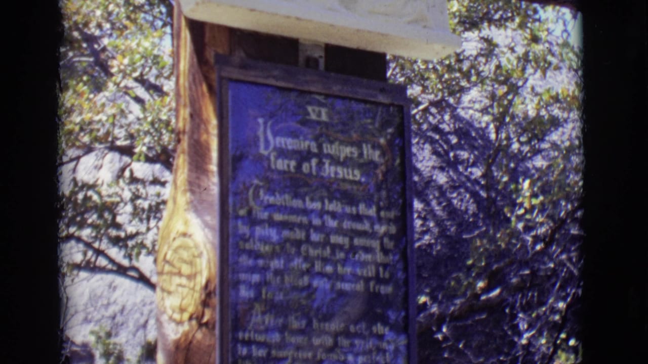 1965 San Luis, Colorado frame with a blue Stations of the Cross plaque, carved hanging ornament, wooden support, and scrubby trees.
