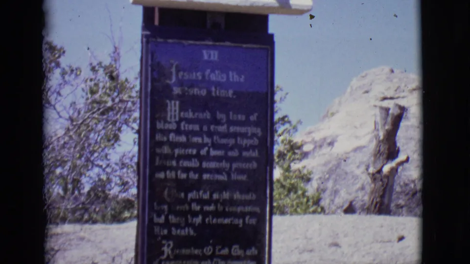 1965 San Luis, Colorado 8mm still showing a Station VII plaque reading partly about Jesus falling, with rock and brush in the background.