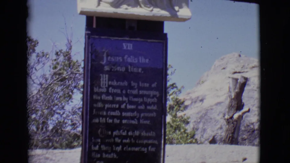 1965 San Luis, Colorado frame showing the Station VII devotional plaque, a partial carved relief above it, trees, and dry rocky terrain.