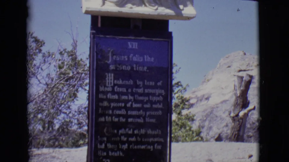 1965 San Luis, Colorado 8mm frame showing a blue Seventh Station plaque beneath a white religious relief sculpture.