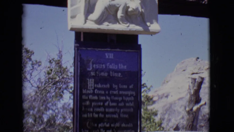 1965 San Luis, Colorado frame showing the Seventh Station plaque and white sculpture with shrubs and rock nearby.