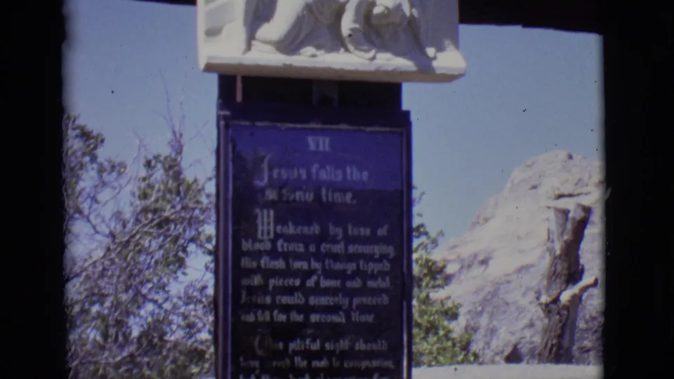 1965 San Luis, Colorado 8mm frame showing a Station VII plaque beneath a white relief at an outdoor religious monument.