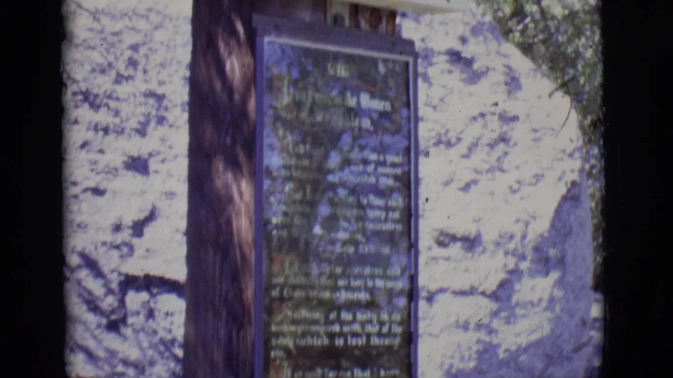 Close 8mm view of a Stations of the Cross plaque on an outdoor monument in San Luis, Colorado, with trees and rock behind it.