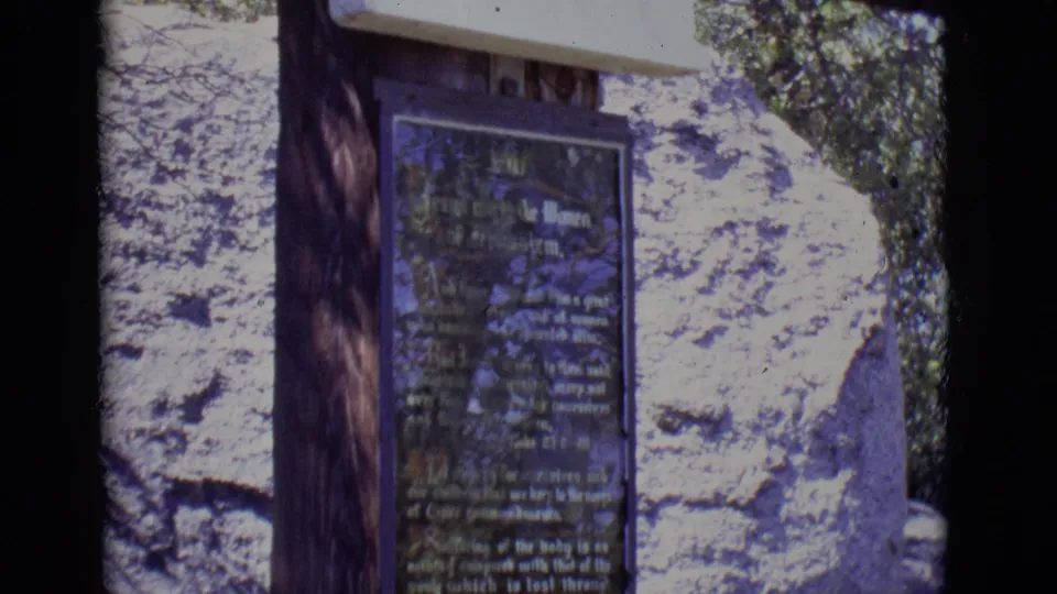 1965 home-movie frame of a religious monument plaque in San Luis, Colorado, partly softened by focus and reflected light.