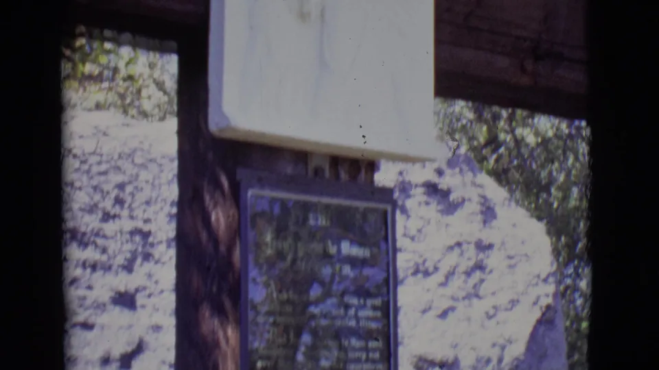 Final 1965 8mm frame showing the religious plaque below a white relief, with San Luis, Colorado rocks and trees in view.