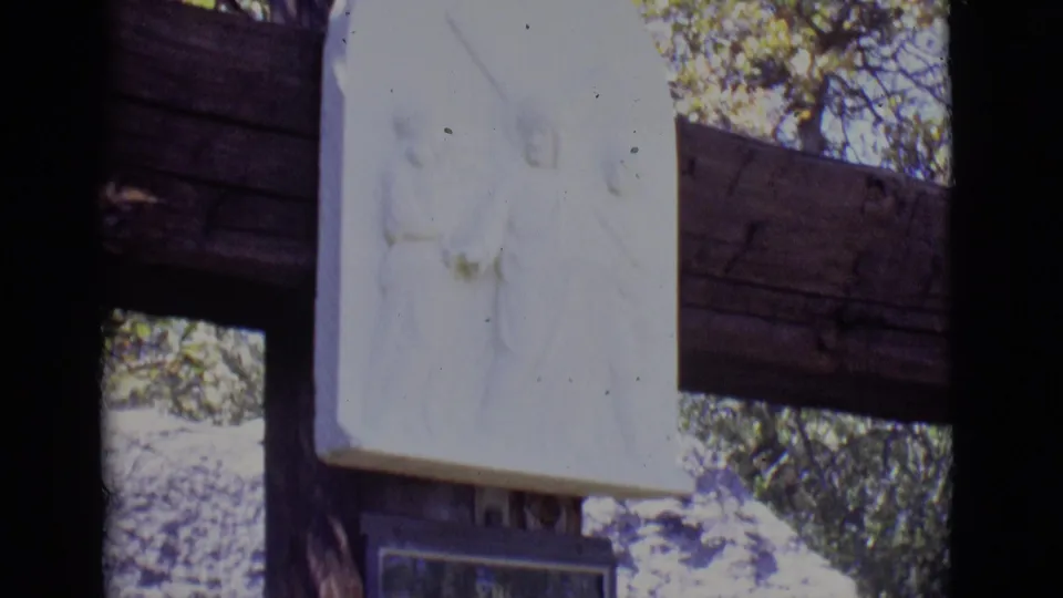 1965 San Luis, Colorado home-movie frame showing a white Stations of the Cross relief above a dark plaque among trees.