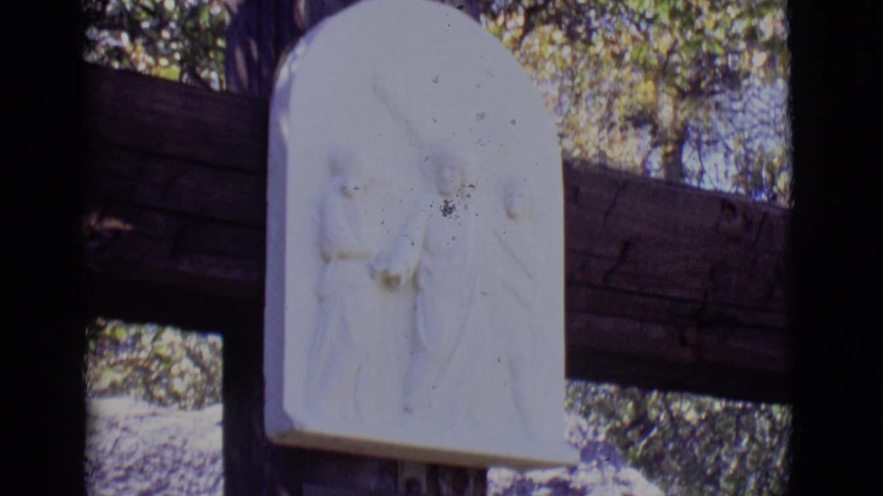 The camera frames the upper portion of the outdoor devotional station, with the white relief and wooden supports prominent.