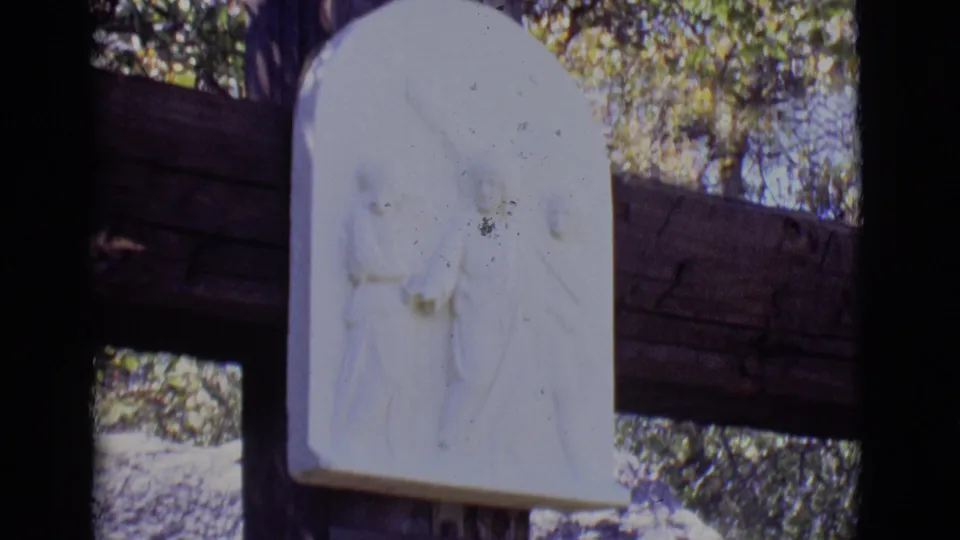 The camera frames the upper portion of the outdoor devotional station, with the white relief and wooden supports prominent.